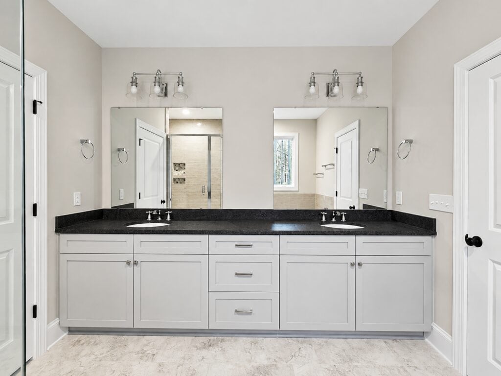 Custom master bathroom design featuring a long dual vanity with gray cabinets, black granite countertops, two mirrors, and a seamless transition to a glass-enclosed shower, by Travars Built Homes.