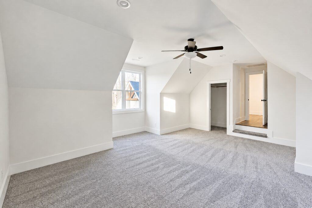 Finished custom home bonus room over the garage featuring plush gray carpet, angled ceilings, a dormer window, and a step-down entry leading to a hallway, built by Travars Built Homes.