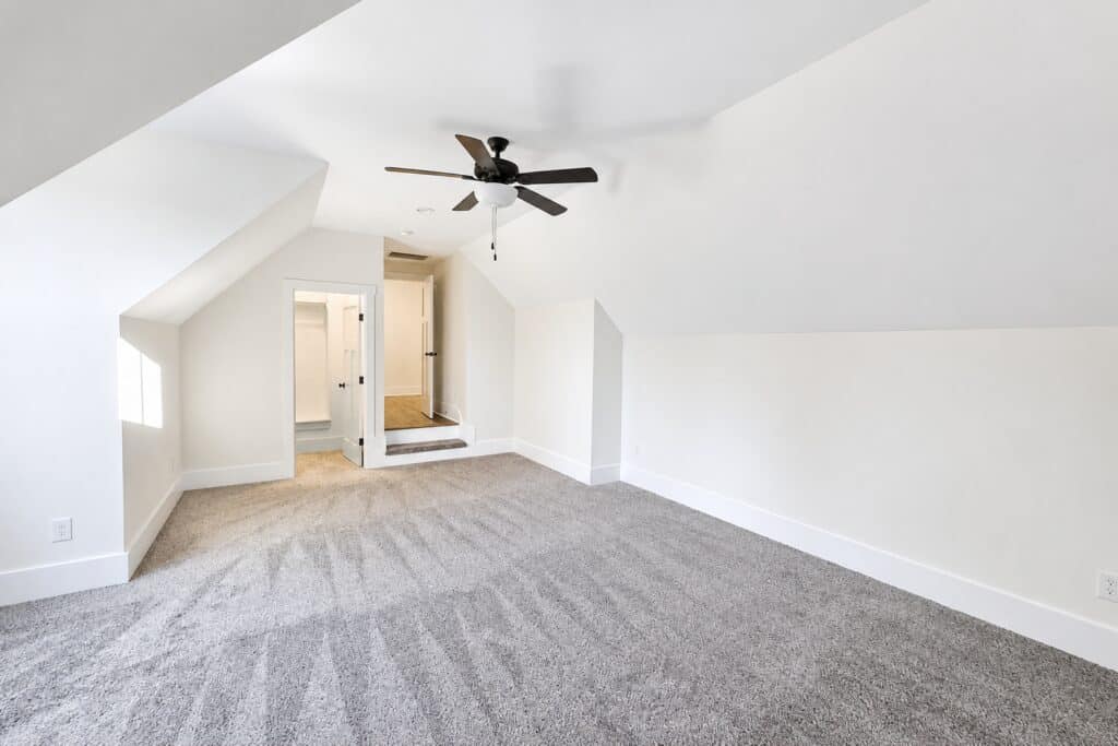 Spacious upstairs bonus room in a custom home showcasing angled ceilings, a modern black ceiling fan, soft carpeting, and steps leading to an adjoining room, crafted by Travars Built Homes.