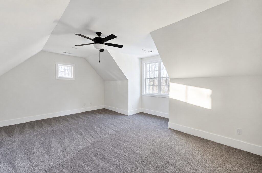 Spacious finished bonus room in a custom home featuring plush gray carpeting, angled ceilings, bright white walls, and a modern ceiling fan, built by Travars Built Homes.
