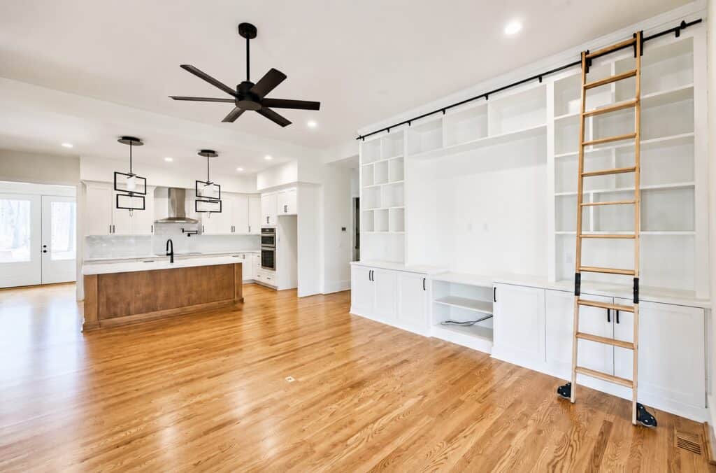Expansive custom living room showcasing warm hardwood floors and a massive built-in white entertainment center featuring multiple shelving cubbies and a rolling wooden library ladder, designed by Travars Built Homes.