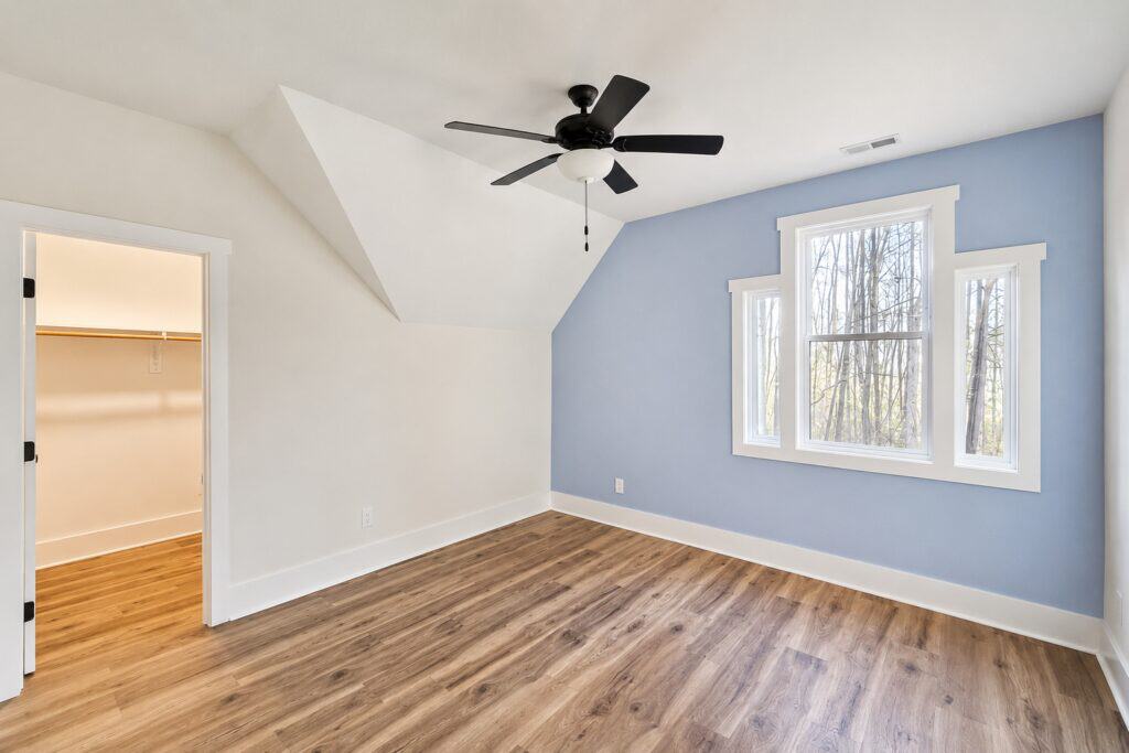 Custom home bonus room or loft space showcasing a light blue accent wall, a unique stepped window design, a modern ceiling fan, and wood-look flooring, built by Travars Built Homes.