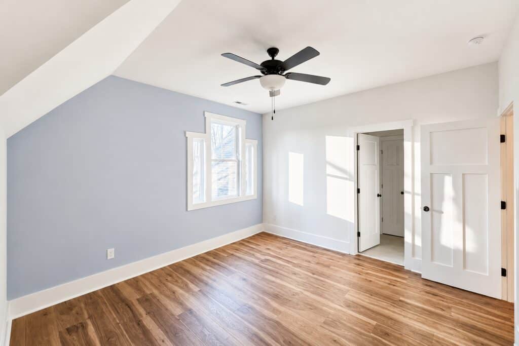 Custom home loft or secondary bedroom featuring warm hardwood flooring, a light blue accent wall, unique stepped window trim, and a modern ceiling fan, built by Travars Built Homes.