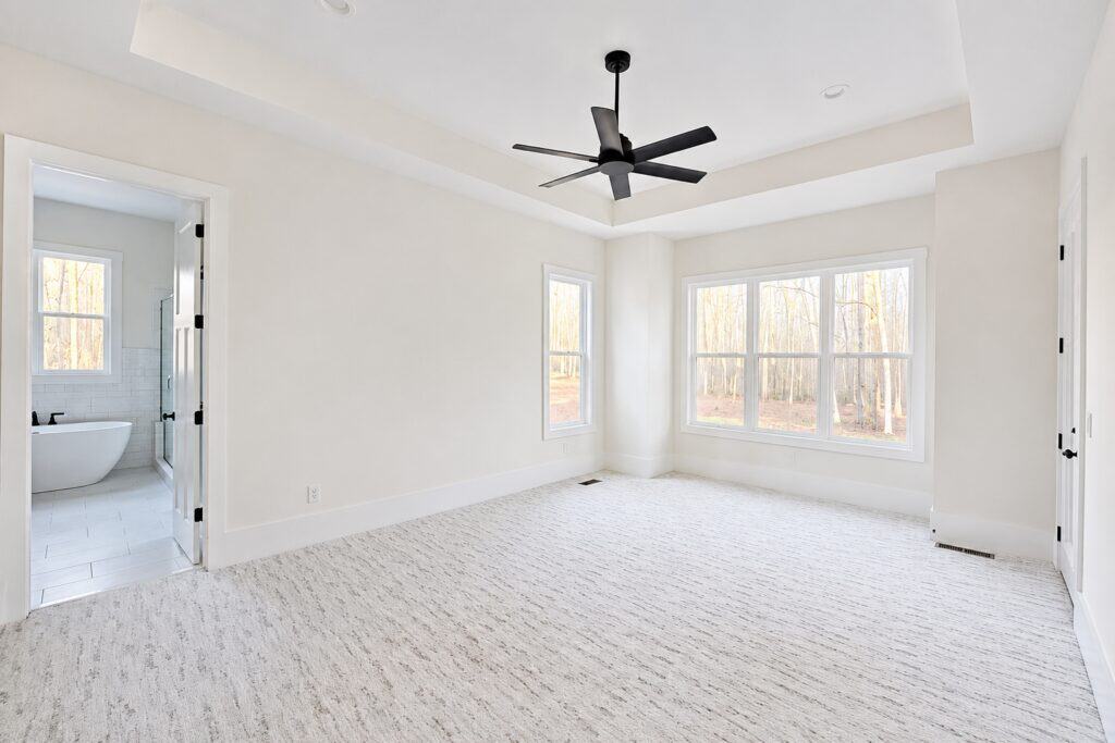 Bright custom master bedroom featuring plush patterned carpeting, an elegant tray ceiling with a modern black fan, large windows, and open double doors leading to a luxury en-suite bathroom, by Travars Built Homes.