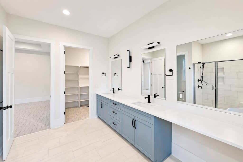 Luxury custom master bathroom showcasing a long light blue dual vanity with white countertops, matte black fixtures, and a large walk-in glass shower, designed by Travars Built Homes.