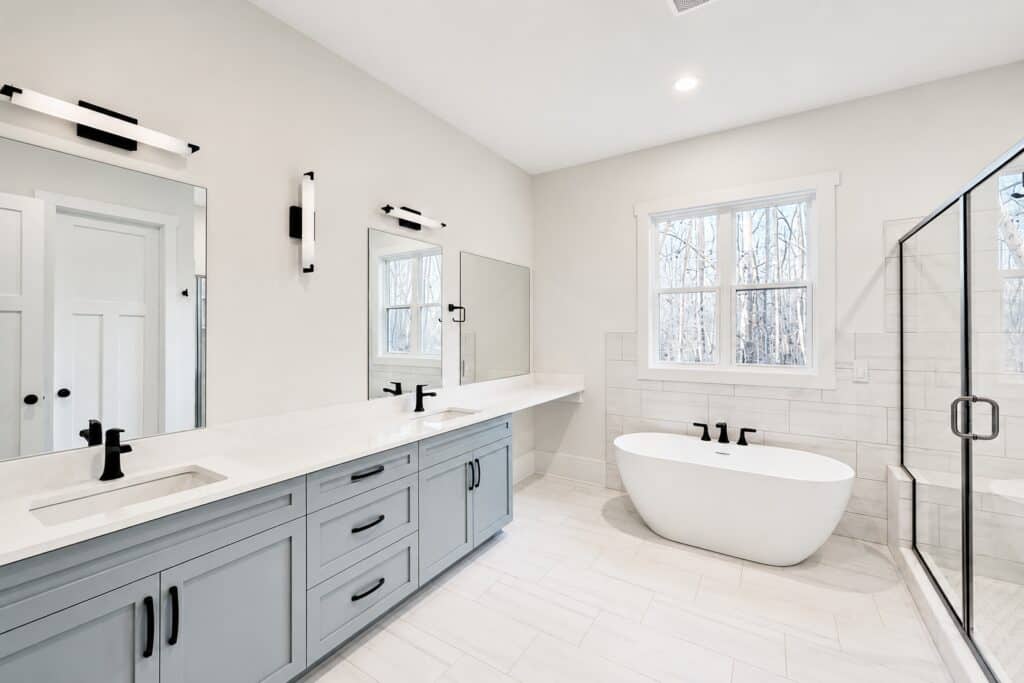 Spacious custom luxury bathroom featuring a light blue double vanity, white quartz countertops, a modern freestanding soaking tub, and a large walk-in glass shower, by Travars Built Homes.