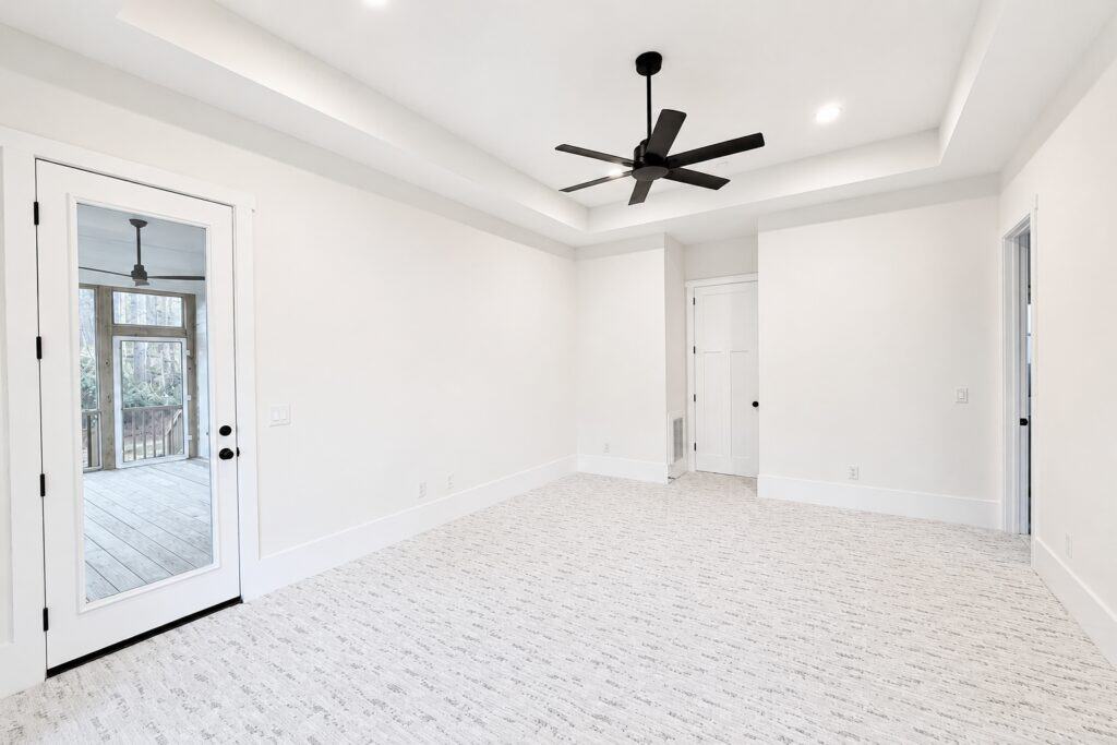 Spacious custom master bedroom showcasing a tray ceiling with a dark modern fan, textured light carpeting, and a glass-paneled exterior door offering private access to a screened-in porch, designed by Travars Built Homes.
