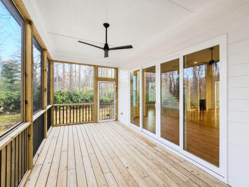 Spacious screened-in porch featuring natural wood decking, a white beadboard ceiling with a modern black fan, black metal railings, and large sliding glass doors leading into the custom home, built by Travars Built Homes.