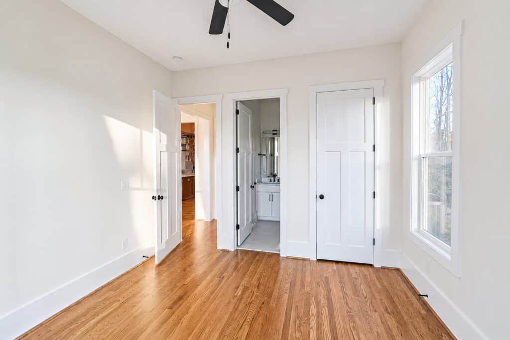 Bright custom secondary bedroom featuring warm hardwood floors, multiple white paneled doors leading to closets and an en-suite bathroom, and ample natural light, crafted by Travars Built Homes.