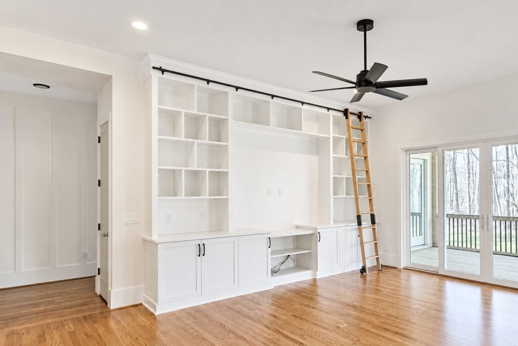 Spacious custom living room showcasing a massive floor-to-ceiling white built-in entertainment center with open shelving, lower cabinets, and a rolling wood library ladder, designed by Travars Built Homes.