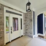 Front porch entry with stone walls, white front door with sidelights and transom, black lantern light, blue siding accents, white arched columns, concrete porch floor, and wooded yard views.