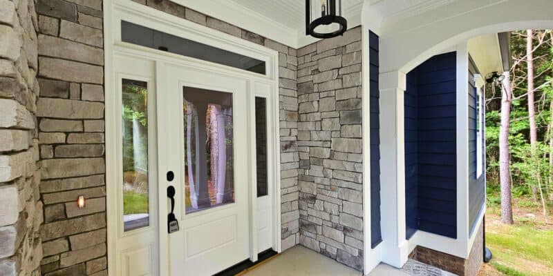 Front porch entry with stone walls, white front door with sidelights and transom, black lantern light, blue siding accents, white arched columns, concrete porch floor, and wooded yard views.
