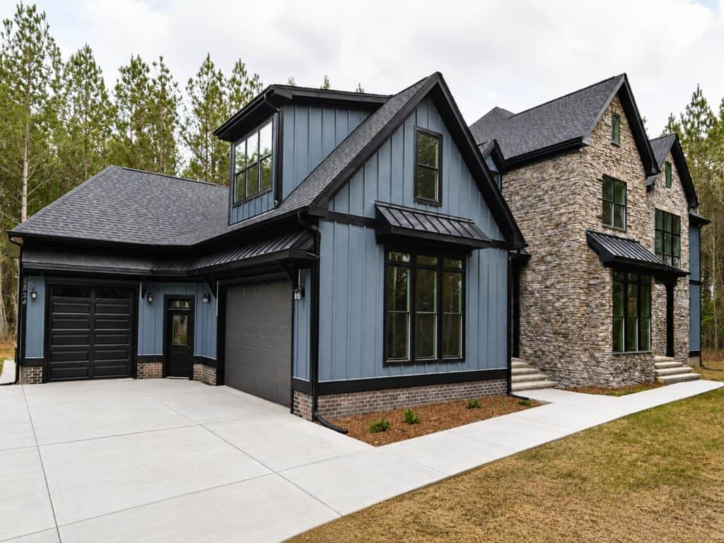 Front exterior view with blue siding, stone accents, black trim, garage bays, concrete driveway, landscaped beds, walkway, multiple gables, and a wooded setting around the home.