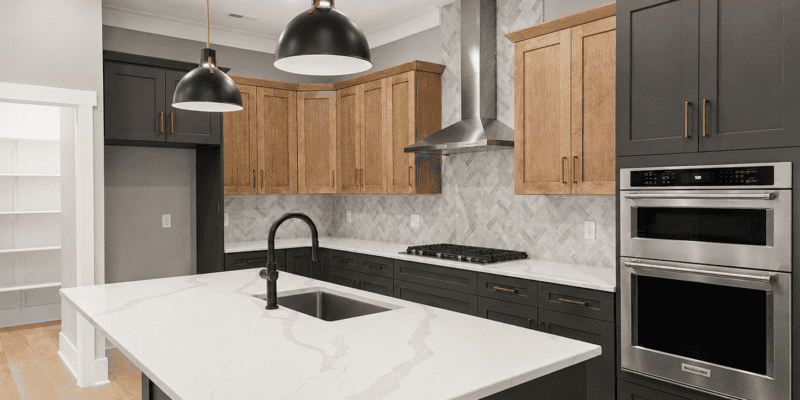 Custom kitchen range wall with black lower cabinets, natural wood upper cabinets, herringbone tile backsplash, gas cooktop, and double ovens by Travars Built Homes.